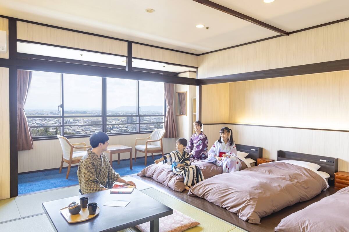 A hotel room with a large window, a bed, a bedside table, and a seating area with chairs and a table.