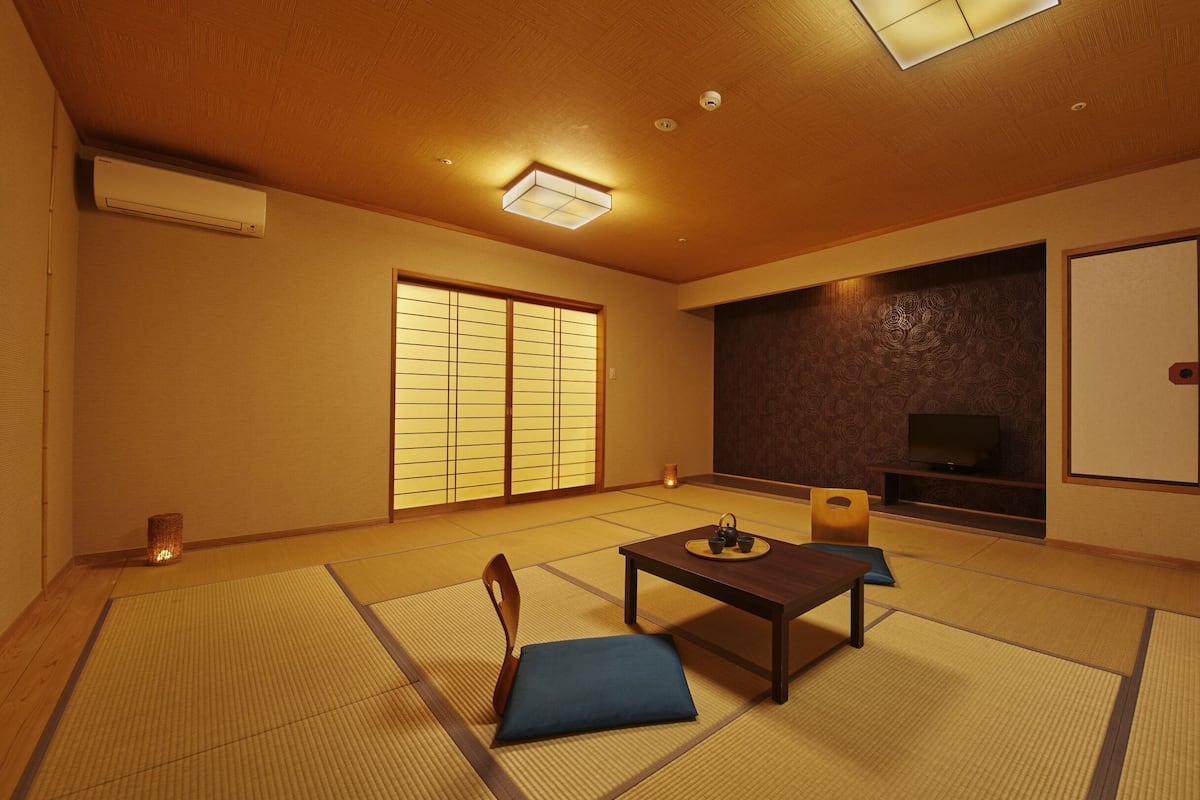 A traditional Japanese room with tatami flooring, a low table with cushions, a television, sliding doors, and a ceiling fan.