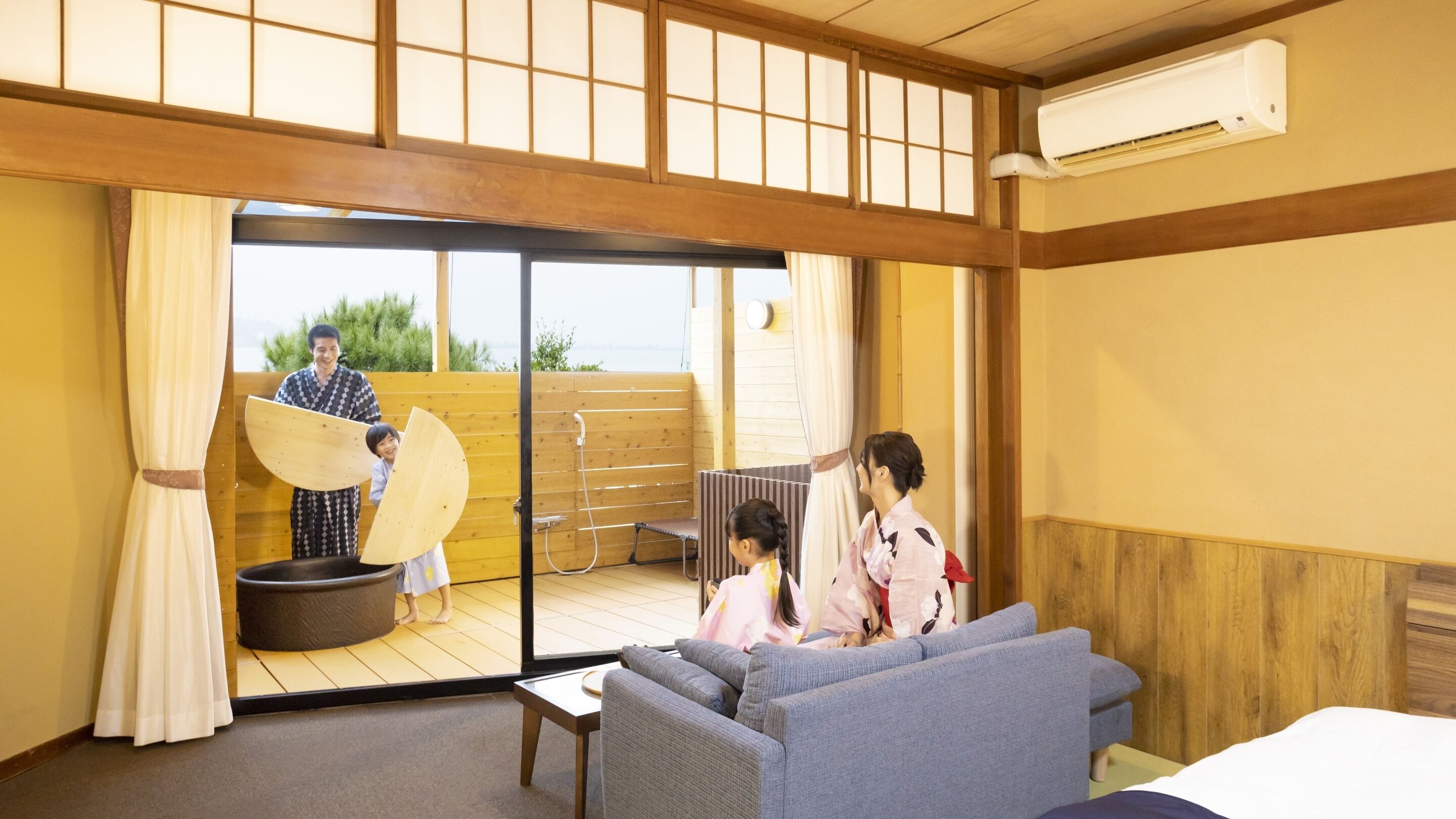 Et traditionelt japansk rum med tatamimåtter, en skydedør af glas der fører til et udendørs område, en sofa og en futon-seng.