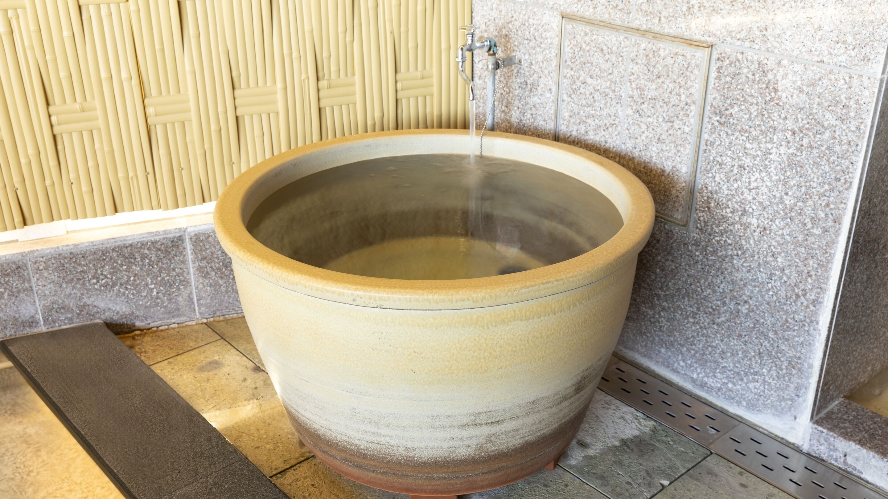 Un grand récipient en céramique rempli d’eau, avec un robinet et un sol carrelé.
