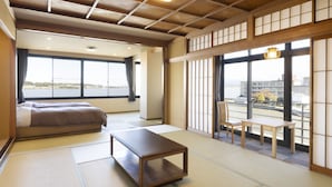 A traditional Japanese-style room with tatami flooring, a low wooden table, a sofa, and a view of a river and buildings. - Ooedo Onsen Monogatari Premium Kaga Maruya (Kaga)