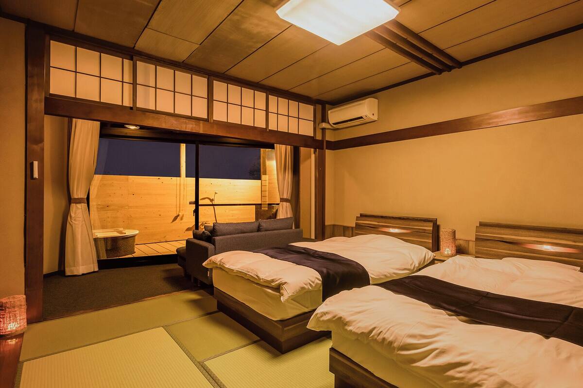 Una habitación tradicional japonesa con suelo de tatami, dos camas con cabeceros de madera, una puerta corrediza con vistas y un equipo de aire acondicionado.