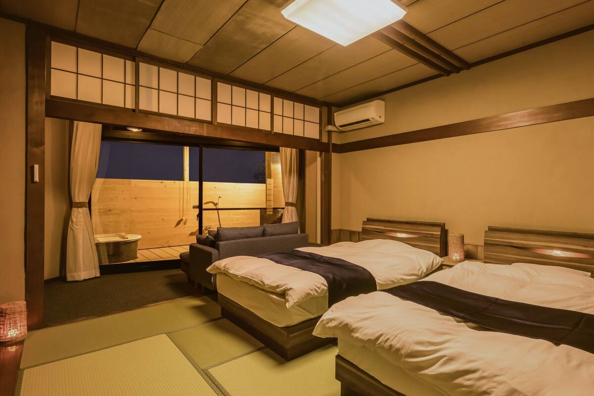 Una habitación tradicional japonesa con suelo de tatami, dos camas de madera con ropa de cama oscura, una puerta corrediza de shoji y un ventilador de techo.