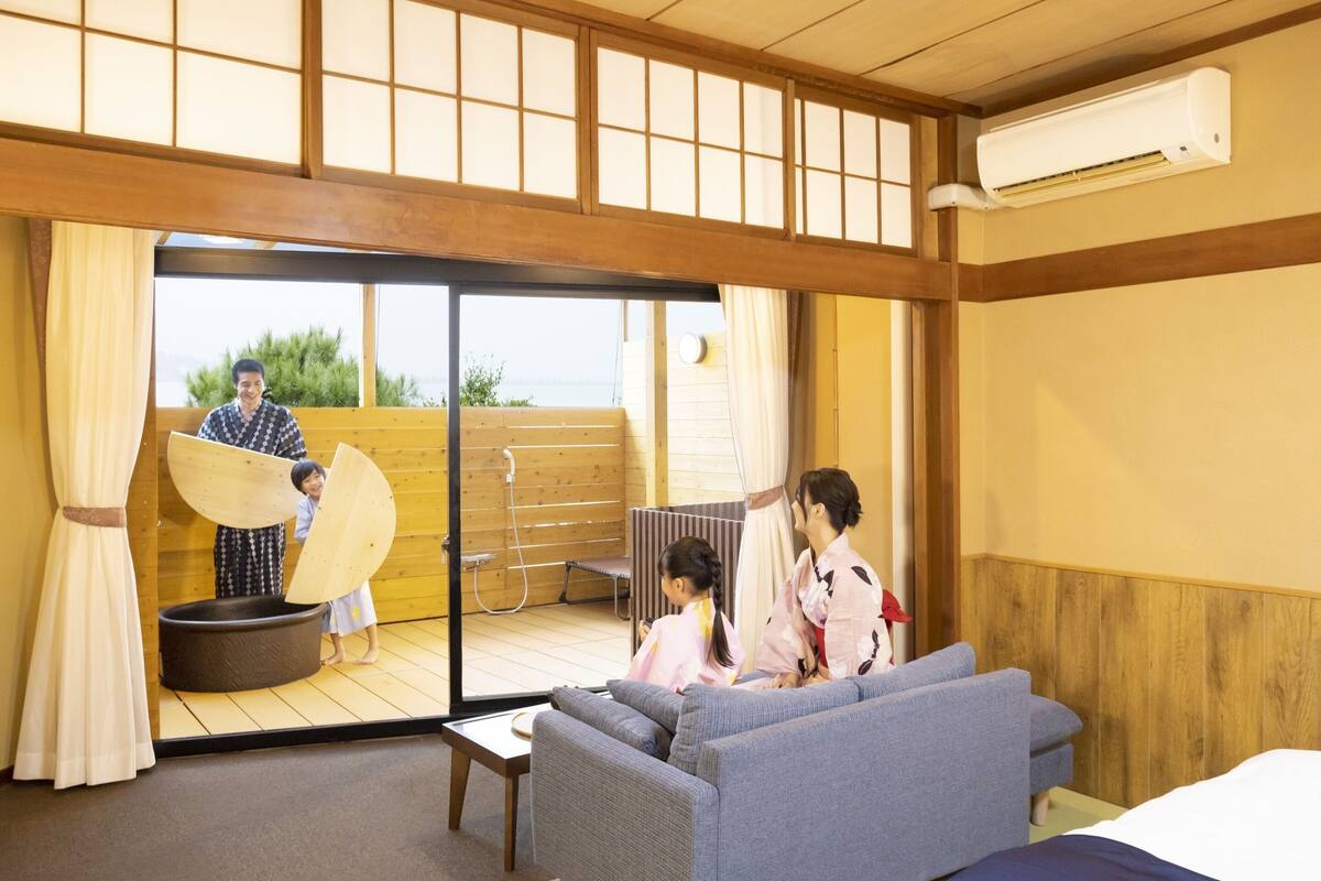 Una habitación japonesa tradicional con suelo de tatami, una puerta corrediza de cristal que da a un balcón, un sofá, una pequeña mesa y un abanico japonés tradicional.