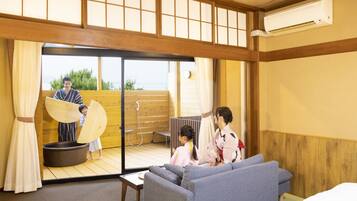 Una habitación japonesa tradicional con suelo de tatami, una puerta corrediza de cristal que da a un balcón, un sofá, una pequeña mesa y un abanico japonés tradicional.