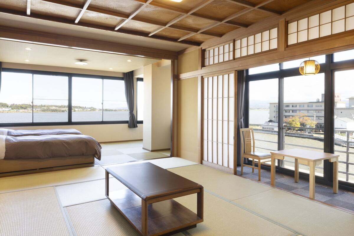 Una habitación de estilo japonés tradicional con suelo de tatami, una mesa de madera baja, un sofá y vistas a un río y edificios.