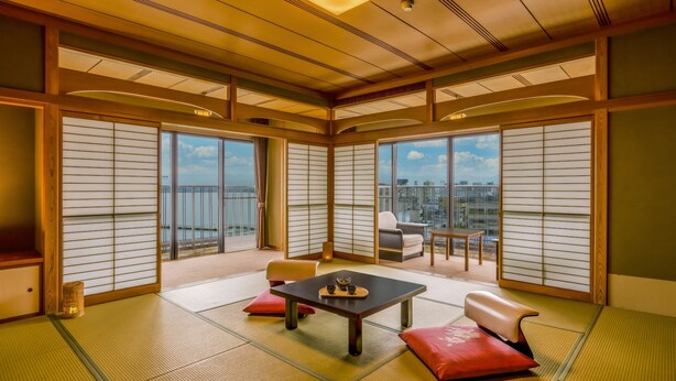A traditional Japanese-style room with tatami flooring, sliding shoji doors, a tatami mat table with a tea set, a sofa, and a view of the city through large windows. - Ooedo Onsen Monogatari Premium Kaga Maruya (Kaga)