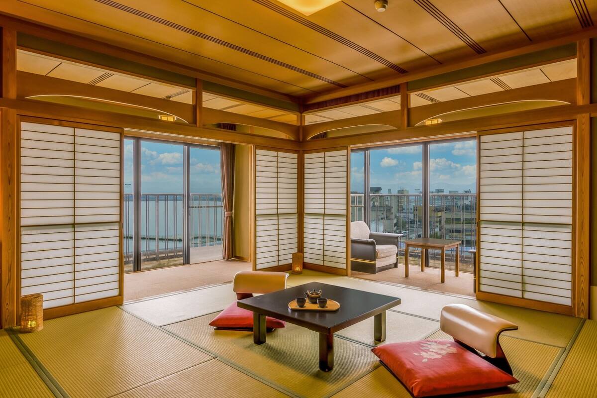 Una habitación tradicional japonesa con suelo de tatami, puertas correderas shoji, una mesa baja de tatami con un set de té, un sofá y vistas a la ciudad a través de grandes ventanales.