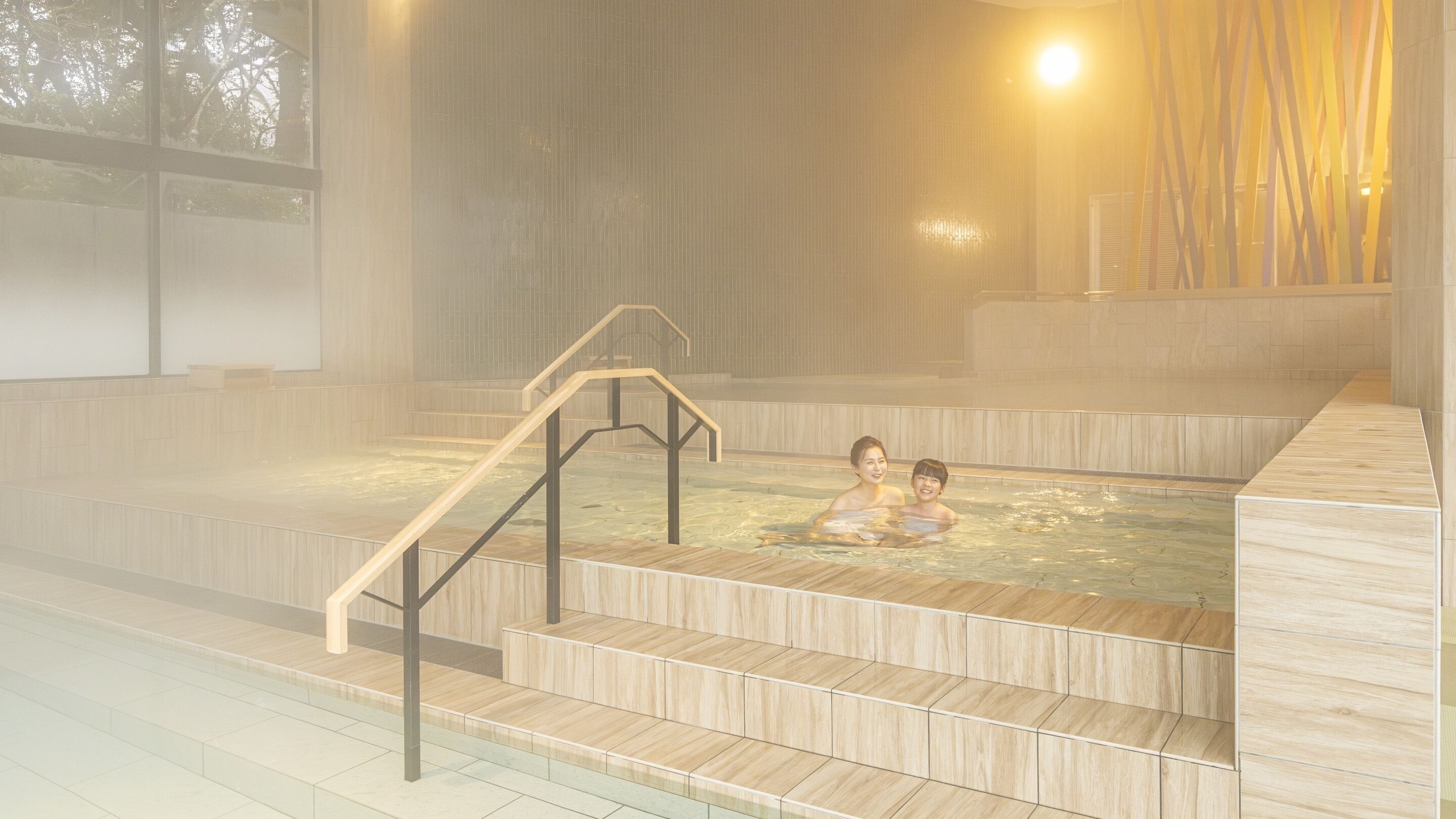 A hot spring bath area with wooden steps leading into the water, a wooden railing, and a view of the outside through large windows.