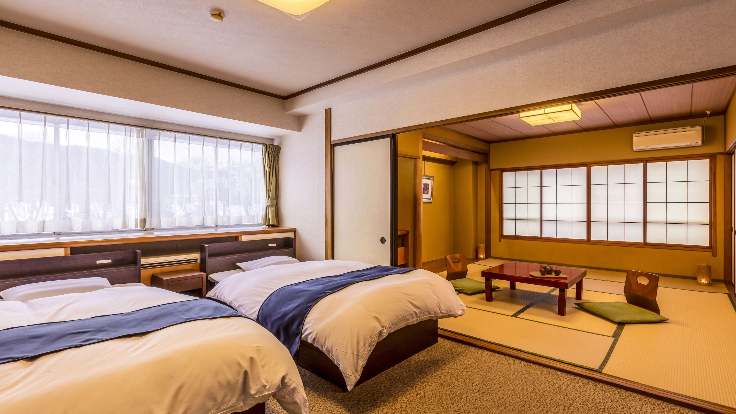Habitación de hotel con dos camas, una zona de estar con mesa y sillas, y vistas a través de un panel shoji.