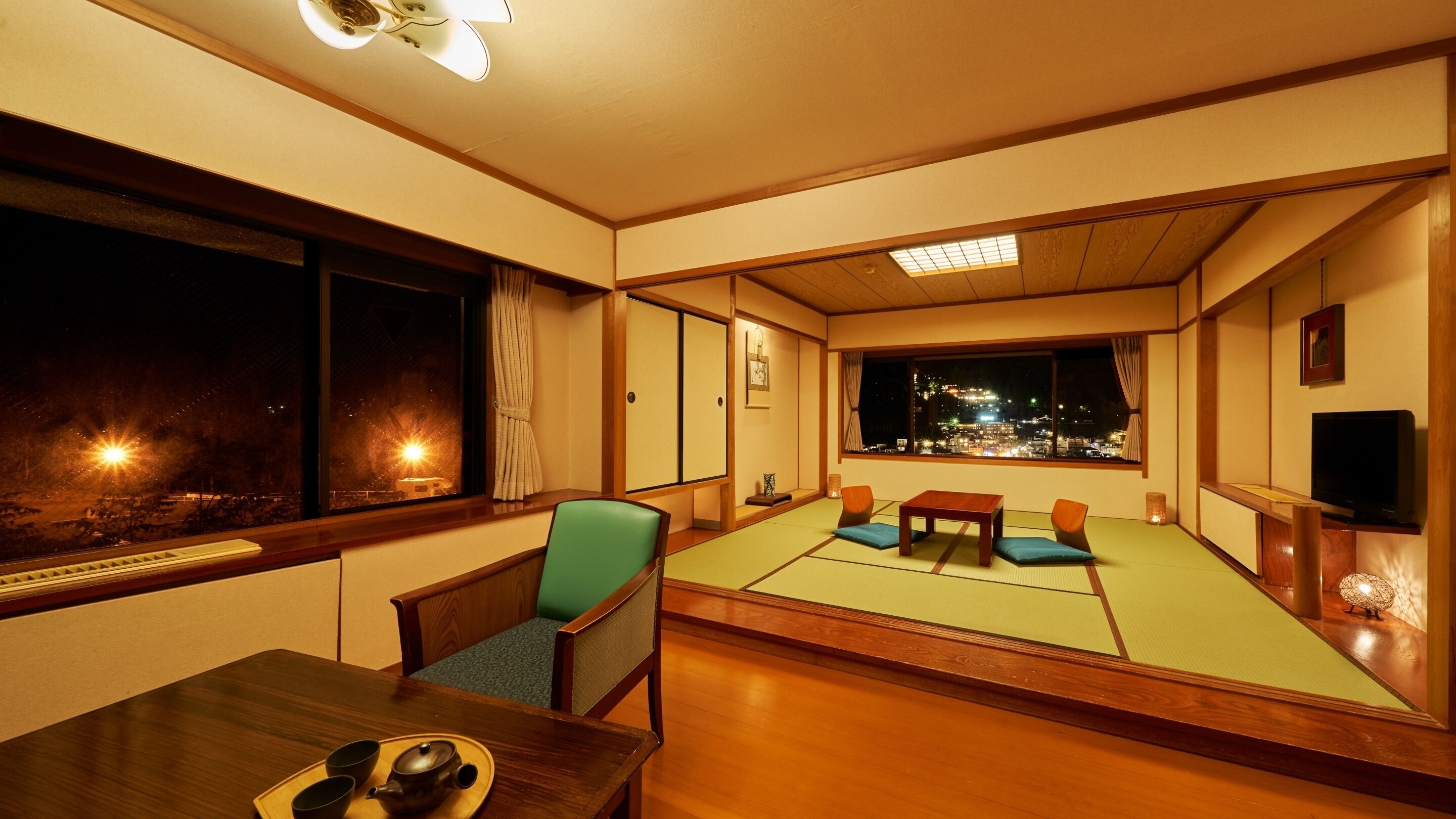 A traditional Japanese room with tatami flooring, a low table with a tea set, a television, and a view of a cityscape at night.