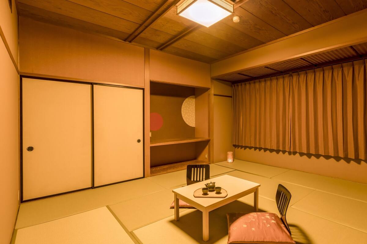 Um quarto japonês tradicional, com tatames, uma porta deslizante, uma mesa com serviço de chá e vista para o exterior através de uma janela com cortinas.