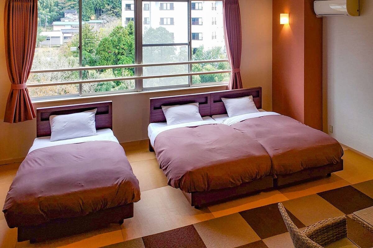 Quarto de hotel com duas camas, uma janela com cortinas, uma mesinha e ar condicionado fixado na parede.
