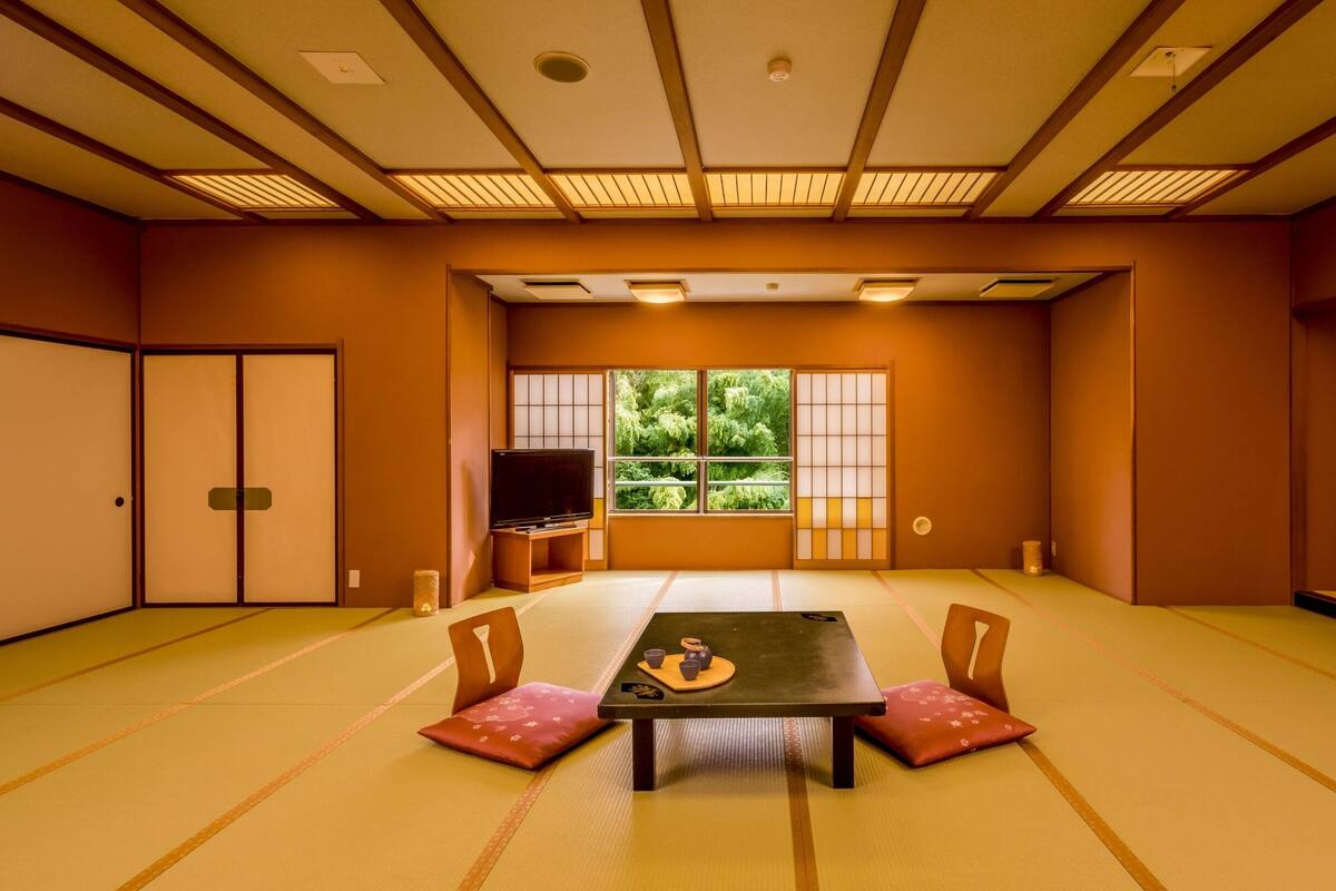Um quarto japonês tradicional com tatame, uma mesa baixa com almofadas, uma televisão e uma grande janela com vista para o verde.