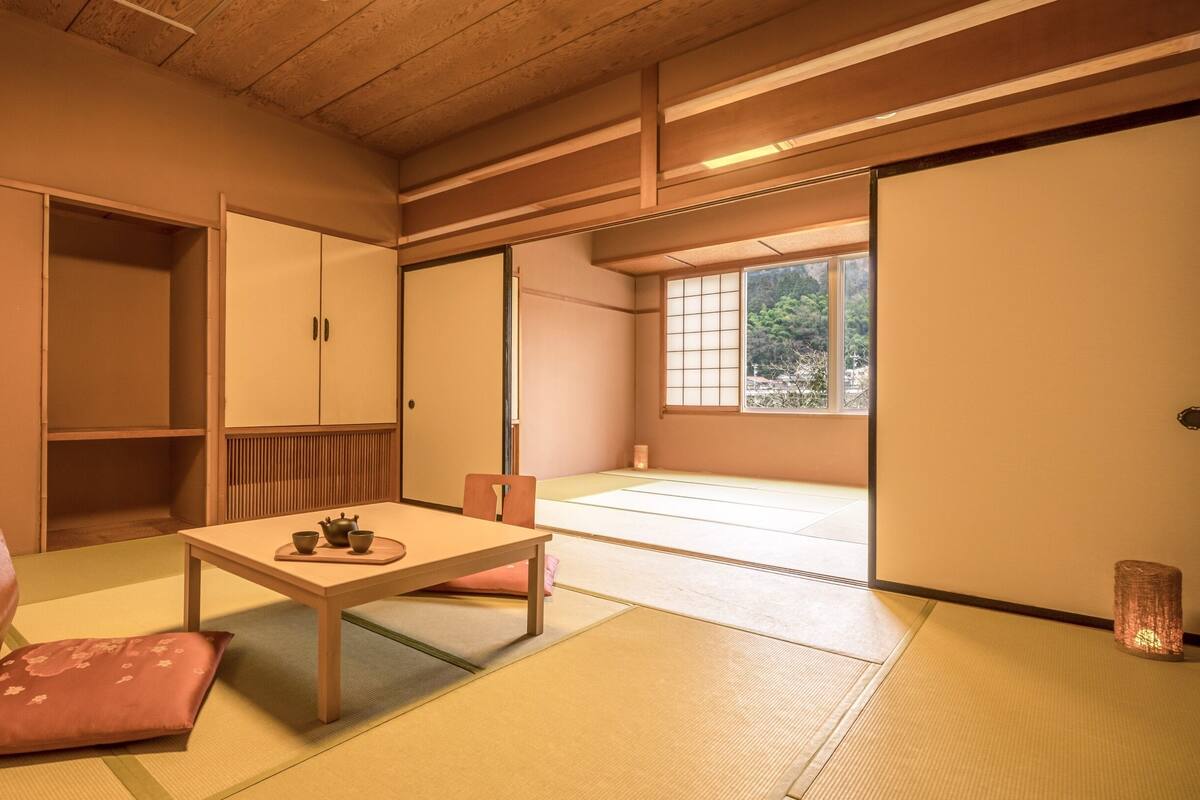 Um quarto japonês tradicional com tatames, uma mesa baixa com um conjunto de chá, uma cadeira de madeira e portas deslizantes que dão para uma varanda com vista para árvores.