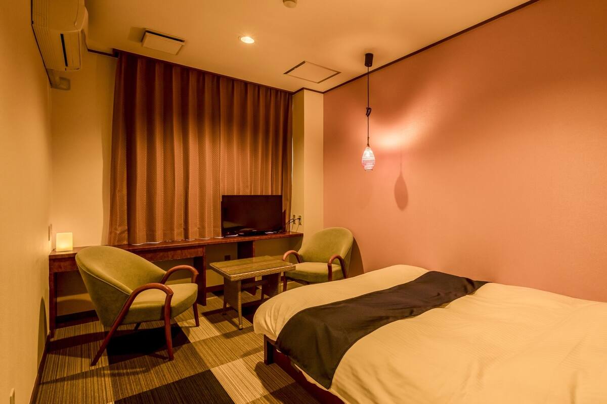 Quarto de hotel com cama, escrivaninha com cadeira, televisão e luminária na parede.