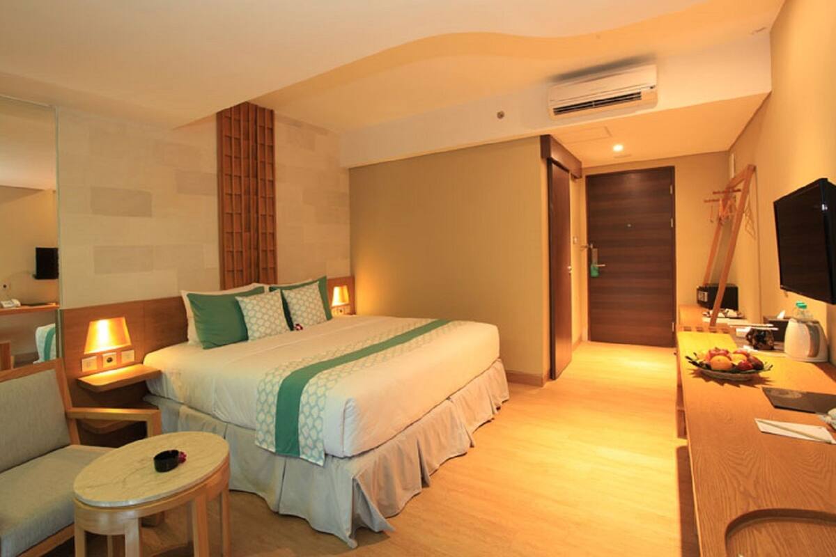 Habitación de hotel con cama, mesitas de noche, escritorio, silla, una pequeña mesa, espejo, televisor y puerta.