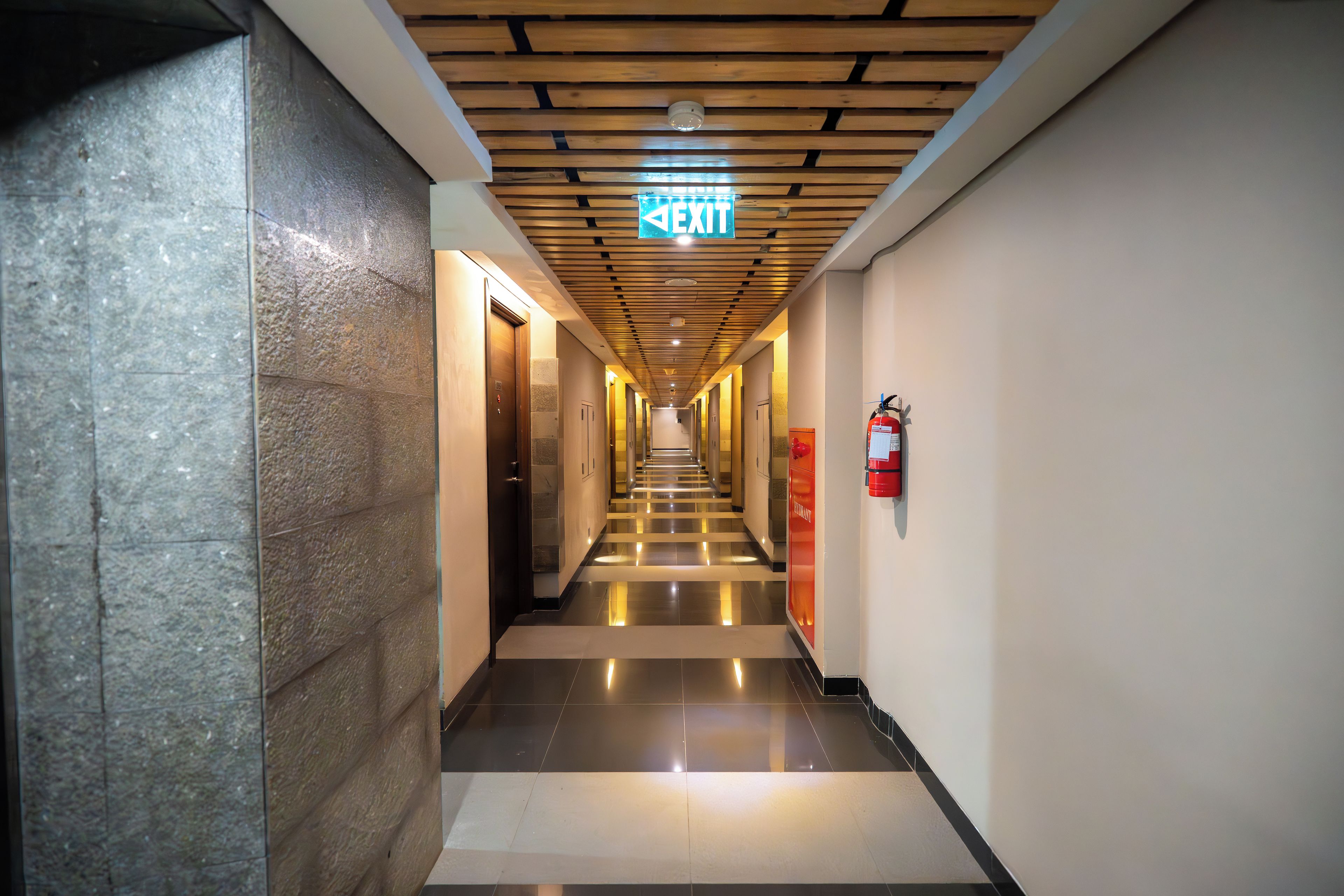 A hotel corridor with a wooden ceiling, a visible exit sign, and a fire extinguisher mounted on the wall.
