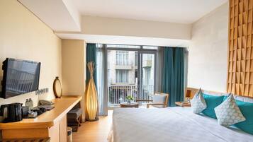 A hotel room with a bed, a desk with a TV, a chair, a small table with a plant, and a balcony with a view of other buildings.