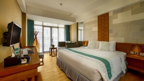 A hotel room with a large bed, a desk with a TV, a chair, a small table with a plant, and a window with curtains. - Bedrock Hotel (Kuta)