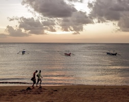 Una playa con tres barcos en el agua y dos personas caminando por la orilla.