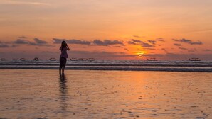 A sunset over a beach with a person standing in the water. Boats are visible in the distance. - Bedrock Hotel (Kuta)