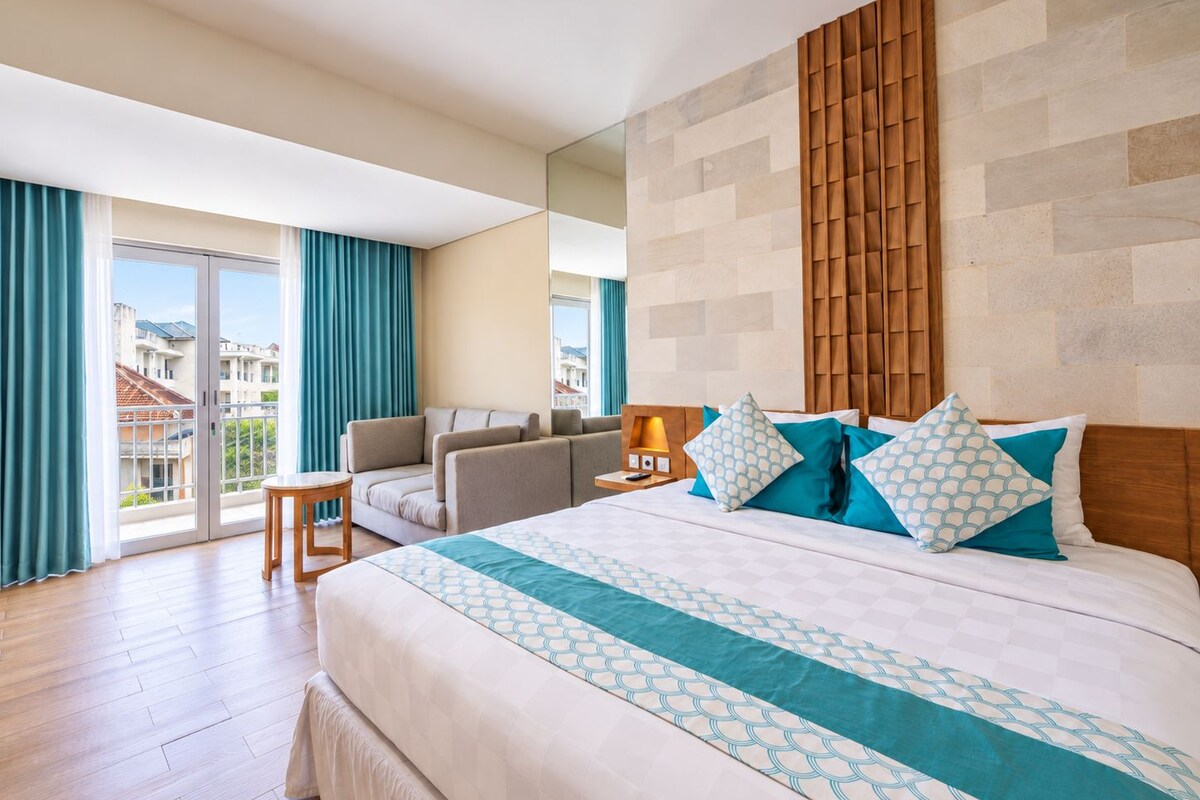 A hotel room with a large bed, a sofa, a small table, and a nightstand with a lamp.