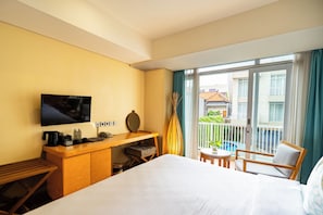 A hotel room with a bed, a desk with a TV, a small table, and a balcony with a view of buildings. - Bedrock Hotel (Kuta)