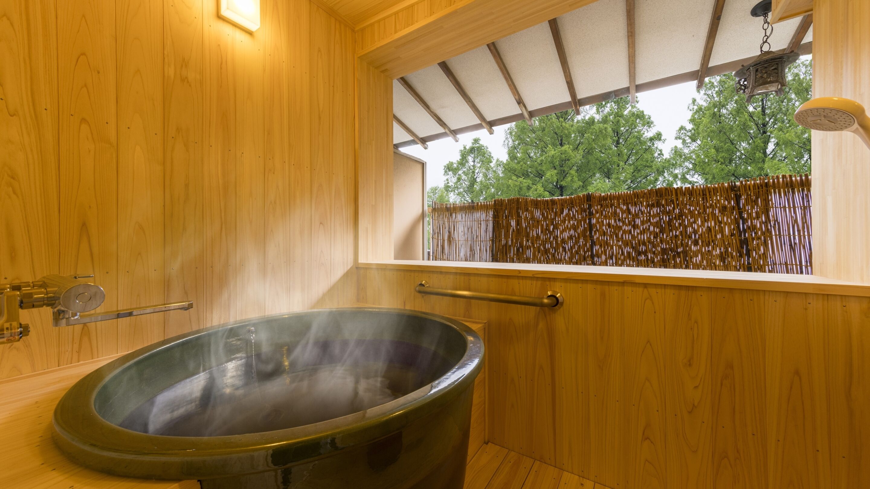 Una sauna de madera con un amplio baño de vapor, una ventana que da a vistas de árboles y una valla de bambú en el exterior.