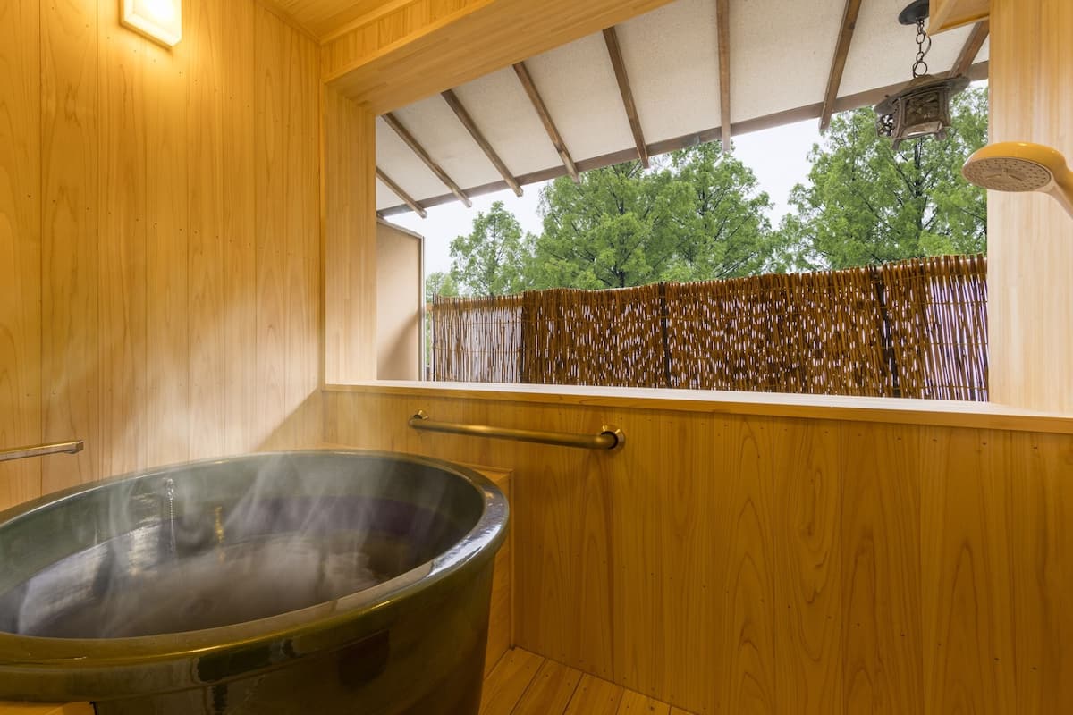 A wooden sauna with a large steam bath, a window with a view of trees, and a bamboo fence outside.