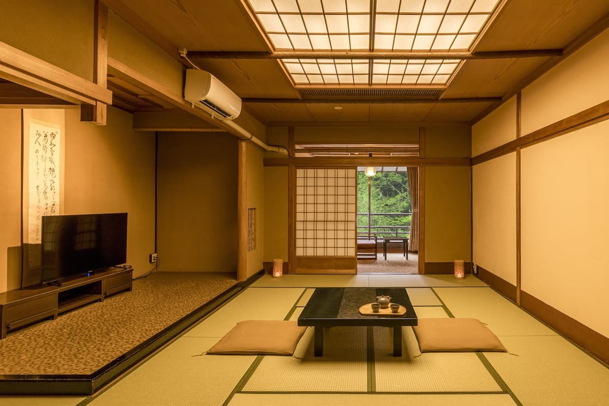 A traditional Japanese room with tatami flooring, a low table with cushions, a television, and a sliding door leading to a balcony.