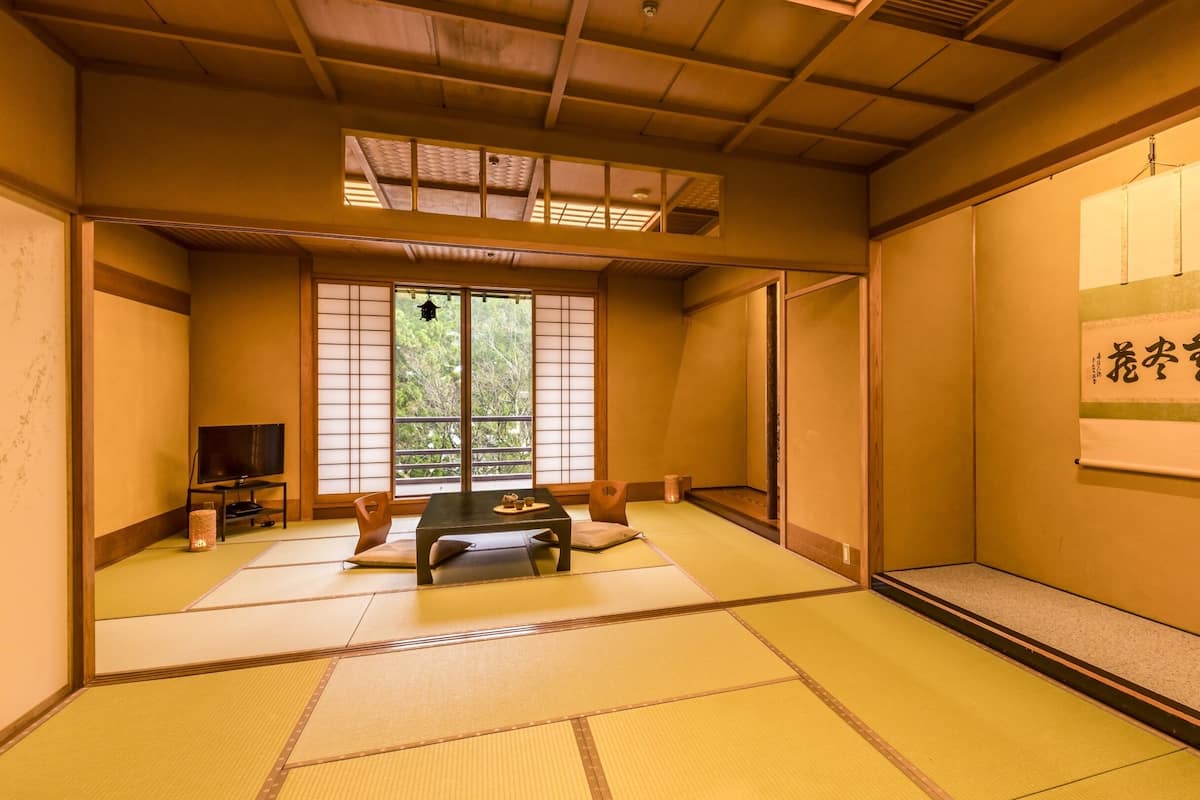 A traditional Japanese room with tatami flooring, a tatami mat table, a low wooden desk with a television, and a sliding door leading to a balcony with a view of trees.
