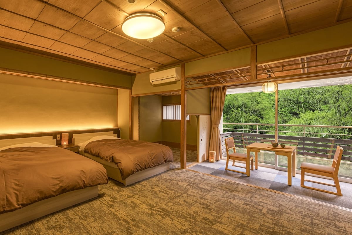 A hotel room with two beds, a small table, a chair, and a balcony with a view of greenery.
