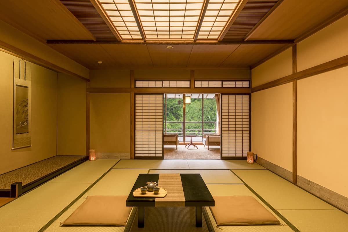 A traditional Japanese room with tatami flooring, a low table with a tea set, sliding doors, and a view of greenery outside.