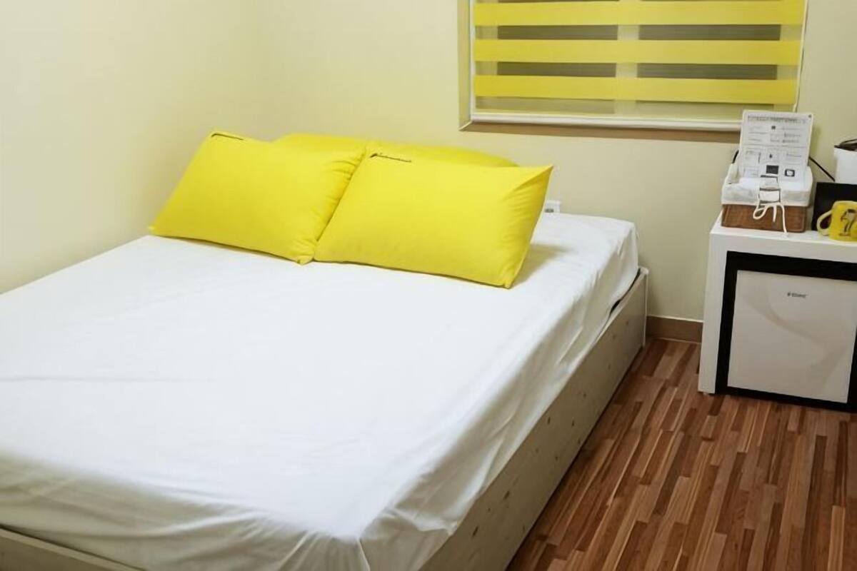 Een bed met witte beddengoed en twee felle gele kussens, een klein nachtkastje met een lamp, en een raam met gele jaloezieën.