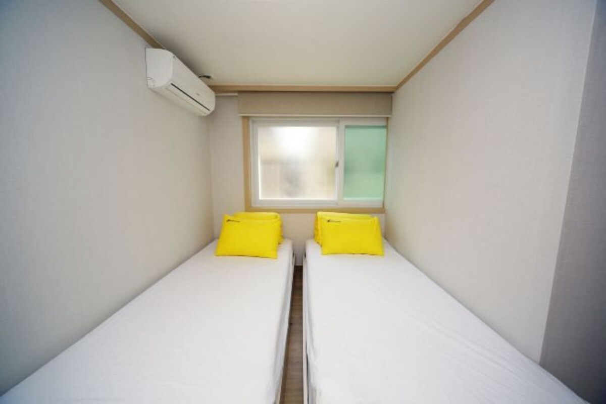 A small, clean room with two single beds, a window with frosted glass, and an air conditioning unit on the wall.