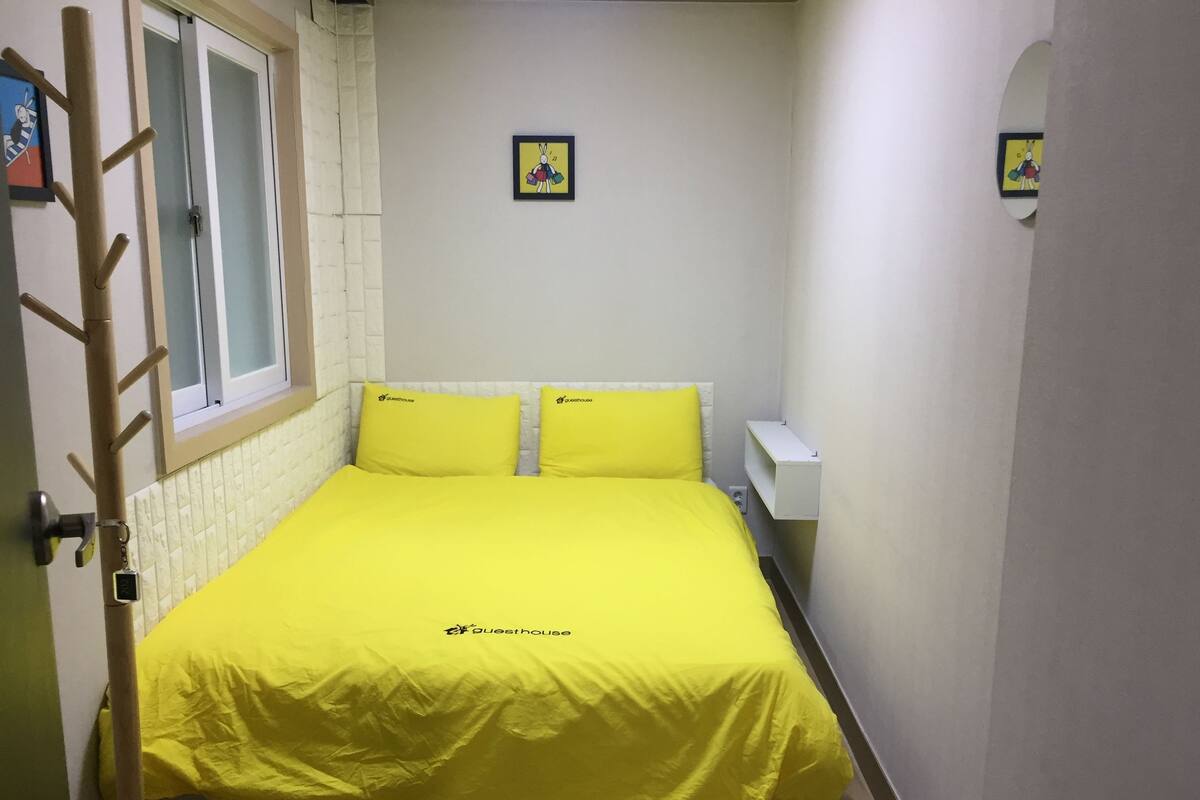 Een kleine, smalle kamer met een bed met geel beddengoed, een raam met uitzicht en een klein schilderijtje aan de muur.