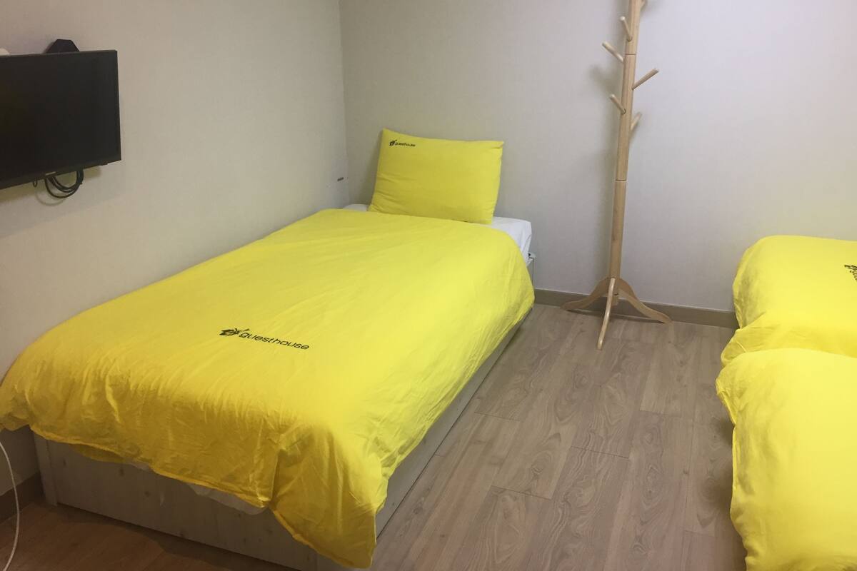 Een kamer met een bed bekleed met gele lakens, een houten kapstok en een flatscreen televisie aan de muur.