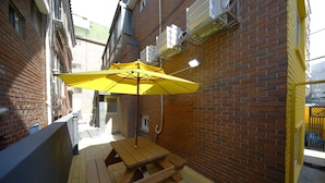 A wooden deck with a picnic table and a yellow umbrella, surrounded by brick walls and air conditioning units. - 24 Guesthouse Seoul Cheongryangri (Seoul)