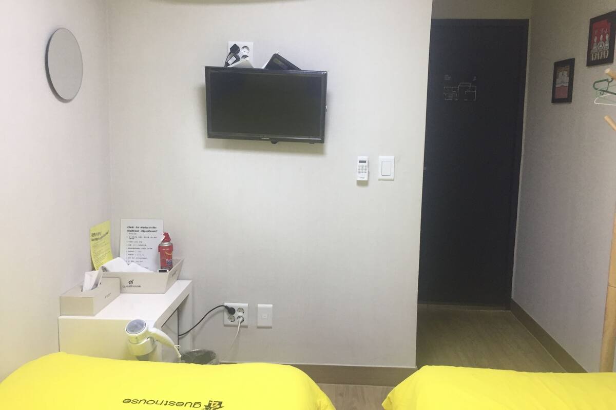 Een hotelkamer met een bed, een aan de muur bevestigde televisie, een klein bureau met een telefoon en een beker, en een deur die naar een andere kamer leidt.
