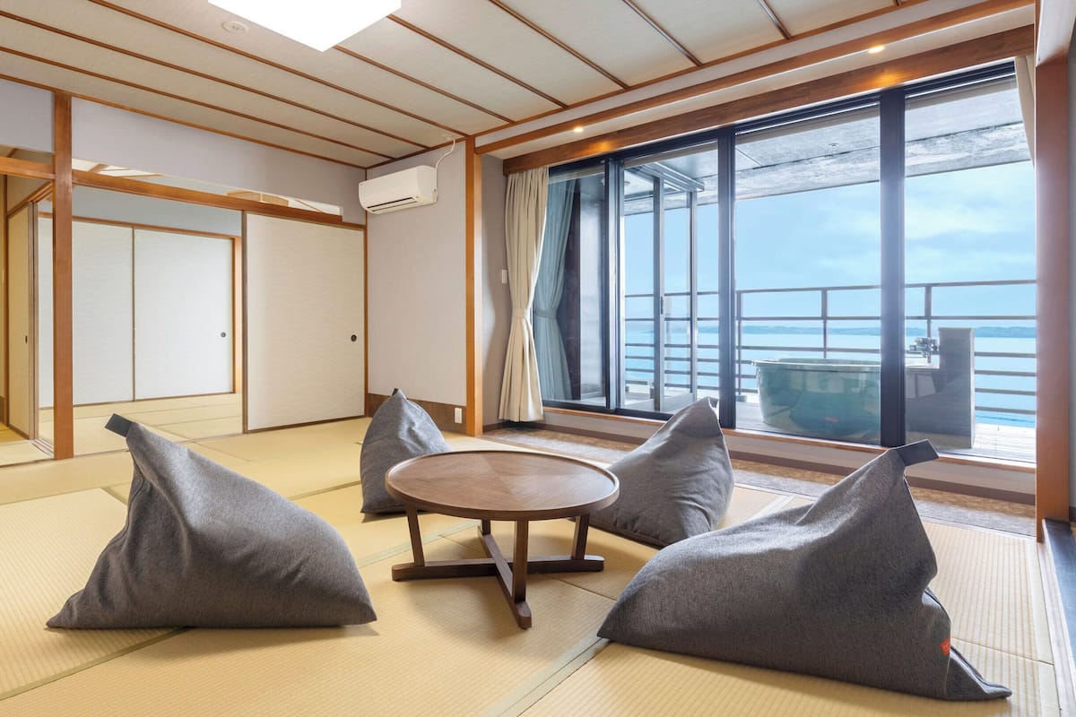 Une pièce avec un sol recouvert de tatamis, une table ronde et des coussins, de grandes fenêtres donnant sur la mer et une porte coulissante.