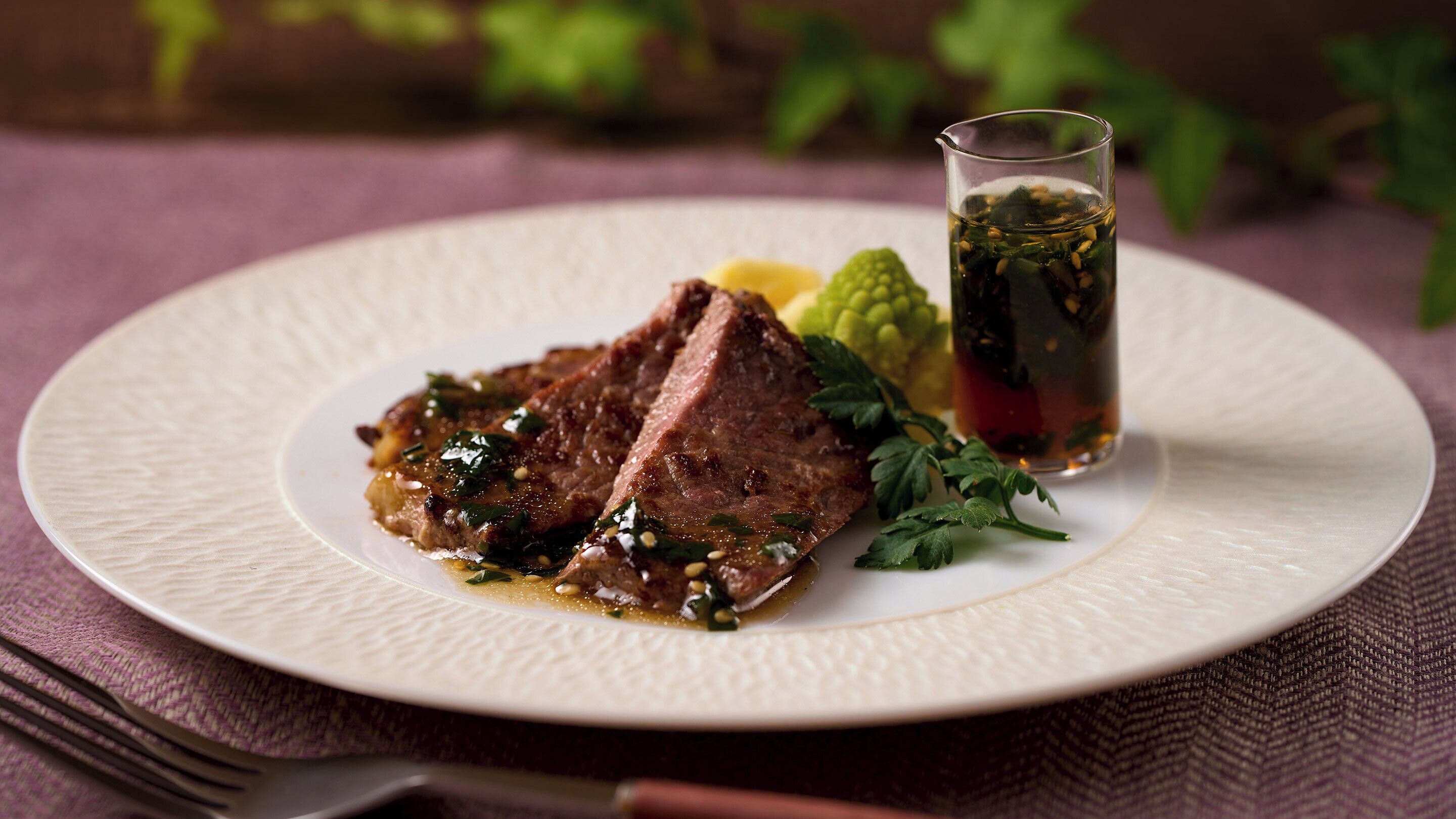 A plate with a steak and a glass of beverage on a textured tablecloth.