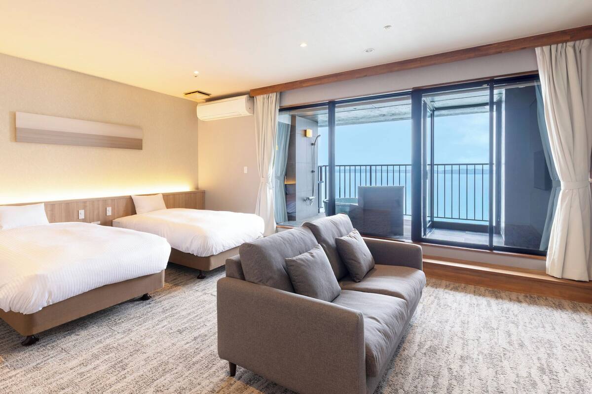 Une chambre d’hôtel avec deux lits, un canapé, une grande fenêtre donnant sur un beau paysage et un balcon.