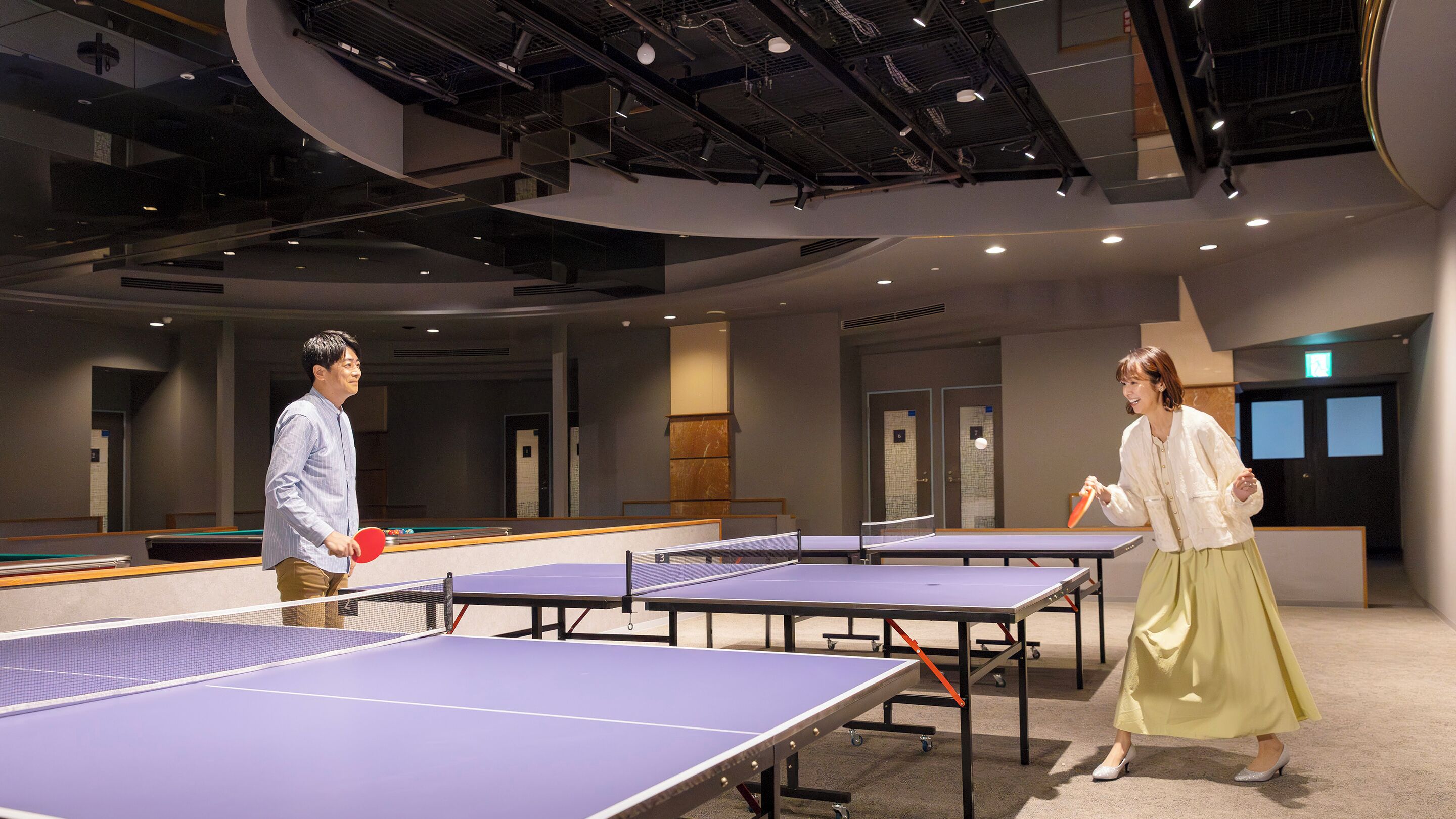 Una stanza con diversi tavoli da ping pong, soffitto alto con travi a vista e arredamento moderno.