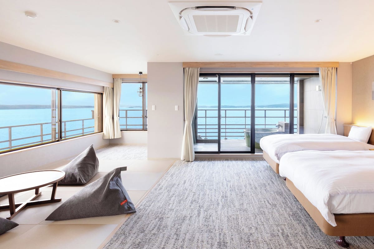 Une chambre d’hôtel moderne avec un grand lit, une petite table, deux chaises, une vue sur la mer grâce à une porte-fenêtre coulissante et un climatiseur au plafond.