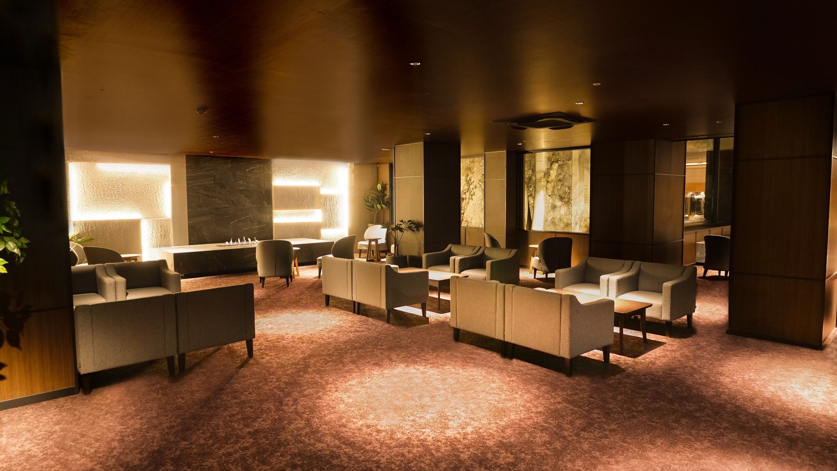 A modern lounge area with sofas and armchairs arranged around a central fireplace, with a stone wall and large windows in the background.