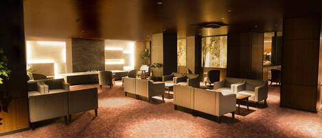 A modern lounge area with sofas and armchairs arranged around a central fireplace, with a stone wall and large windows in the background.