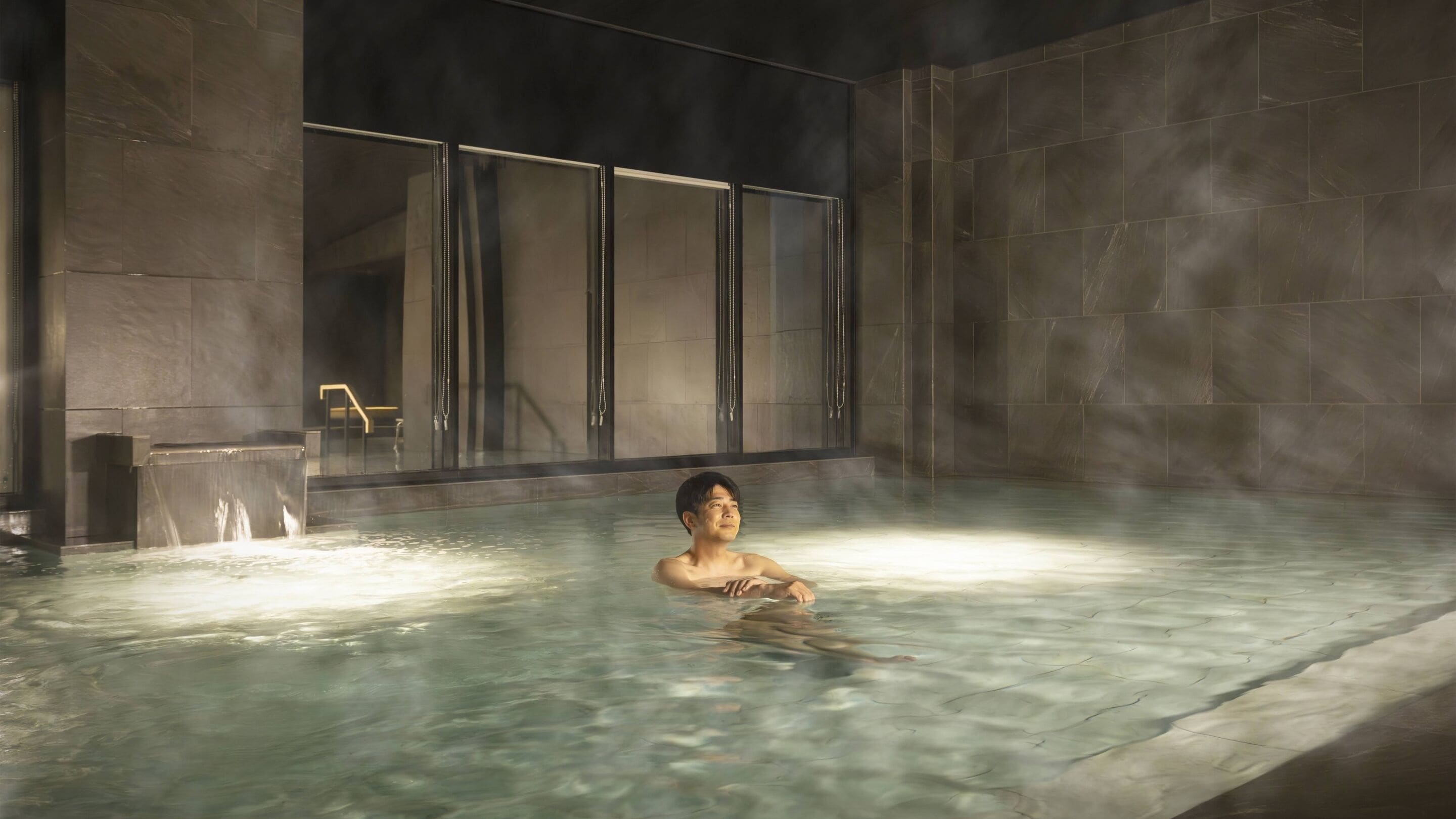 Une personne dans un bain à remous, de la vapeur s'élevant autour d'elle, le tout entouré de murs en pierre et séparé par une paroi de verre.