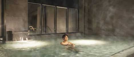 Une personne dans un bain à remous, de la vapeur s'élevant autour d'elle, le tout entouré de murs en pierre et séparé par une paroi de verre.
