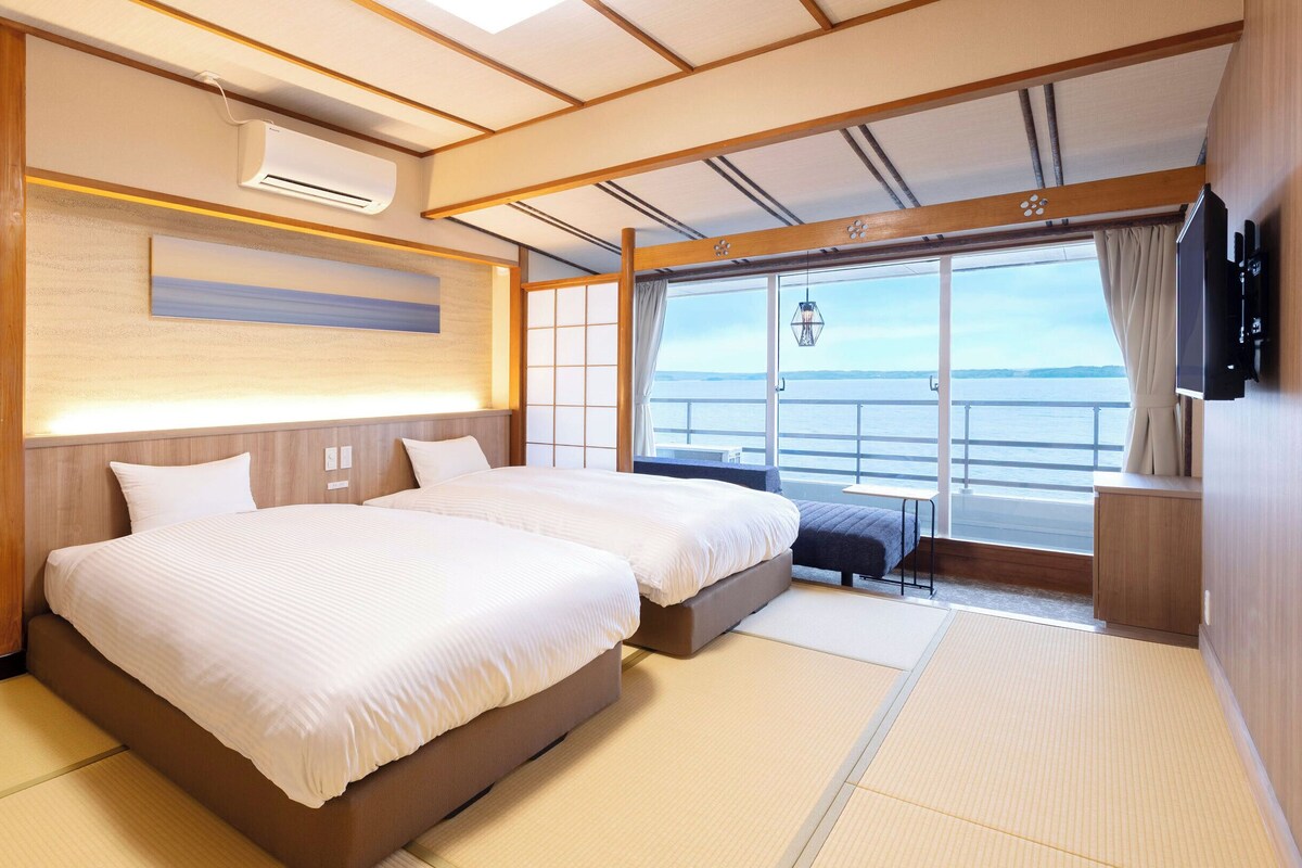 Une chambre d’hôtel avec deux lits, une télévision, une fenêtre donnant sur la mer et un appareil de climatisation.